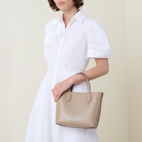 Model carrying Kate Spade Bleecker Medium Leather Crossbody Bag in taupe with gold-tone hardware and top handles, styled with a white midi dress