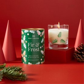 Wax Lyrical Fir and Frost Candle in glass vessel with holly decoration, beside festive green tube packaging on red background