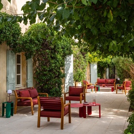 FERMOB Somerset Bench in Chili red with teak frame, styled in a sun-dappled courtyard with matching garden seating and lush greenery