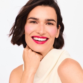 Model wearing Trinny London Lip2Cheek Colour in Milly, a vivid pink-red shade, applied to lips and cheeks, wearing a cream sleeveless top