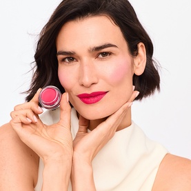 Model with Trinny London Lip2Cheek Colour in Milly, a pink-red shade, holding open pot to cheek, wearing cream top