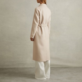 Model wearing Reiss Maeve Double-Breasted Trench Coat in beige, longline wool-blend with self-fabric belt, styled with wide-leg white trousers