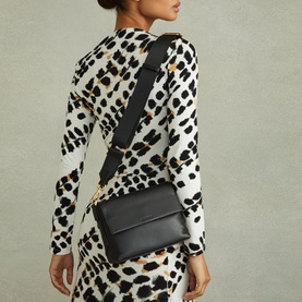 Model carrying Reiss Selena Logo Crossbody Bag in black leather with gold-tone hardware and wide crossbody strap, styled with leopard-print dress