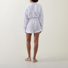 Model wearing Sea Level Sunset Beach Shorts in white cotton cheesecloth, high-rise with drawstring waist, styled with matching top and gold sandals