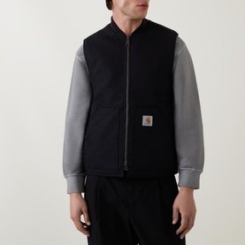 Model wearing Carhartt Logo Vest Gilet in black, sleeveless with zip front and logo patch, styled with grey long-sleeve top and black trousers