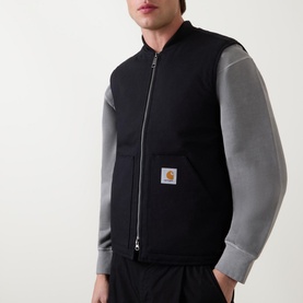Model wearing Carhartt Logo Vest Gilet in black, sleeveless with kangaroo pockets and logo patch, styled over grey long-sleeve top