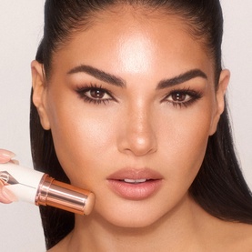 Model with highlighter stick applied to cheekbone, rose gold packaging, glowing bronzed complexion finish