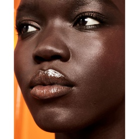 Model wearing FENTY Gloss Bomb Oil in Coppa Cookie, a warm glossy lip look with subtle shimmer, against an orange background