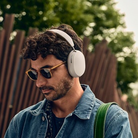 Model wearing Sonos Ace Headphones in white, paired with denim jacket and sunglasses, outdoors with green bag strap visible.