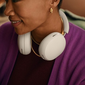 Model with Sonos Ace Headphones in white, worn around neck, gold-tone ear cups visible, styled with purple jacket and gold hoop earrings