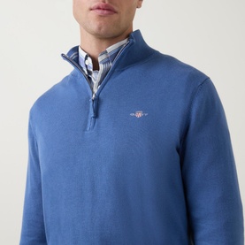 Model wearing Gant Shield Half-Zip Logo Sweater in blue, featuring funnel neckline and embroidered logo, styled with checked shirt underneath