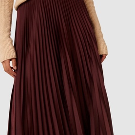 Model wearing Forever New Estelle High-Rise Pleated Skirt in burgundy, satin pleated fabrication, styled with a cream knit jumper