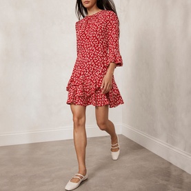 Model wearing Mint Velvet Bow Print Ruffle Mini Dress in red, three-quarter sleeves, ruffle hem, styled with white flat shoes