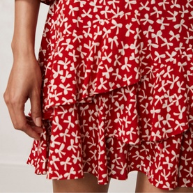 Model wearing Mint Velvet Bow Print Ruffle Mini Dress in red with cream bow print, close-up of tiered ruffle skirt detail