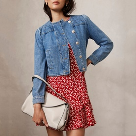 Model wearing Mint Velvet Bow Print Ruffle Mini Dress in red, styled with a denim jacket and white shoulder bag