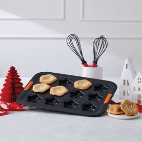Le Creuset 12-Cup Non-Stick Star Tray 40cm in slate grey with orange handles, filled with star-shaped bakes, styled in a festive Christmas setting