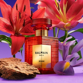 Balmain Rouge Eau de Parfum in red and gold bottle, styled with red lilies, purple iris and driftwood on a violet background.