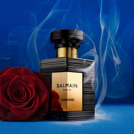 Balmain Carbone Eau de Parfum in black and gold ribbed bottle, styled with a red rose against a blue background with smoke