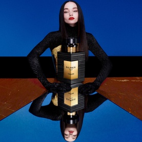 Model with Balmain Carbone Eau de Parfum, black and gold bottle reflected on glossy surface against bold blue backdrop