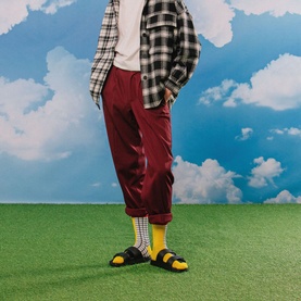 Model with Eat My Socks French Fries Socks in yellow, worn with red trousers, check shirt and yellow sandals on a grass backdrop