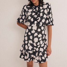 Model wearing Mint Velvet Abstract Tiered Mini Dress in black monochrome floral print, puff sleeves, bow neckline, and tiered skirt