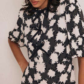 Model wearing Mint Velvet Abstract Tiered Mini Dress in black monochrome print with bow neckline detail and short puff sleeves