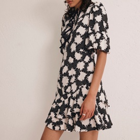 Model wearing Mint Velvet Abstract Tiered Mini Dress in black monochrome floral print with puff sleeves, bow neckline, and tiered skirt