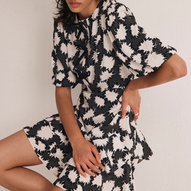 Model wearing Mint Velvet Abstract Tiered Mini Dress in black monochrome floral print with puff sleeves, bow neckline, and tiered skirt