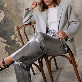 Model wearing Mint Velvet Grey Single-Breasted Blazer with metallic silver trousers and black sandals, seated on wooden chair