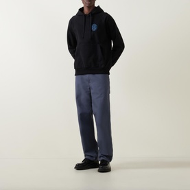 Model wearing Deus Ex Machina Pill Logo Hoodie in black with circular chest logo, styled with blue wide-leg trousers and black loafers