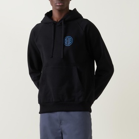 Model wearing Deus Ex Machina Pill Logo Hoodie in black with blue circular chest logo, styled with grey trousers
