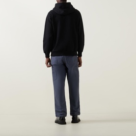 Model wearing Deus Ex Machina Pill Logo Hoodie in black, rear view, styled with navy trousers and black shoes