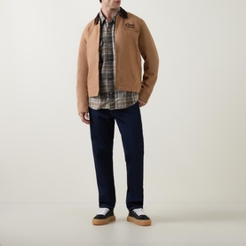 Model wearing Deus Ex Machina Address Barn Jacket in tan with corduroy collar, styled with plaid shirt, navy trousers and trainers