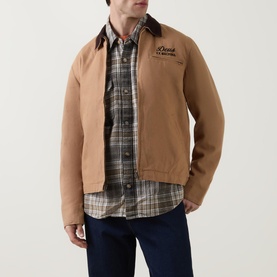 Model wearing Deus Ex Machina Address Barn Jacket in tan with corduroy collar, styled with plaid shirt and navy jeans