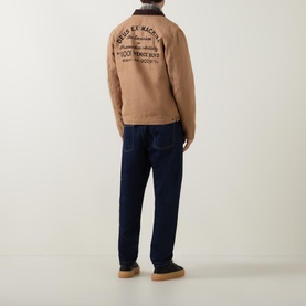 Model wearing Deus Ex Machina Address Barn Jacket in tan canvas, back view showing logo print, corduroy collar, styled with dark navy jeans