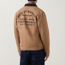 Model wearing Deus Ex Machina Address Barn Jacket in tan, showing embroidered back logo and corduroy collar, styled with dark jeans