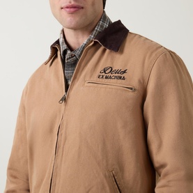 Model wearing Deus Ex Machina Address Barn Jacket in tan, showing embroidered logo and brown corduroy collar, styled with plaid shirt beneath