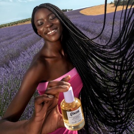 Model with GISOU Honey Infused Hair Perfume in Lavender Berry 50ml, holding bottle aloft in lavender field, wearing pink top