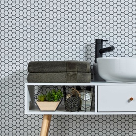 Christys Prism Towel in Tarmac black, folded on a white vanity shelf beside a round basin, small plant, and decorative accessories