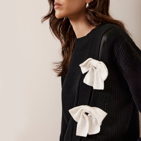 Model wearing Mint Velvet Bow Knit Sweater in black with white oversized bow appliqu&eacute;s, ribbed knit texture, crew neck visible at shoulder
