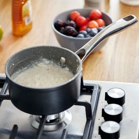Stellar Rocktanium Non-Stick Milk Pan 700ml in black on gas hob with bubbling contents, bowl of fresh fruit in background