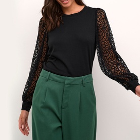 Model wearing Kaffe Becca Lace Top in black with sheer floral lace sleeves and fitted cuffs, styled with green wide-leg trousers