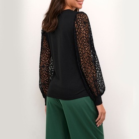 Model wearing Kaffe Becca Lace Top in black with sheer lace long sleeves and fitted cuffs, styled with green wide-leg trousers