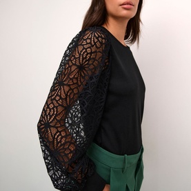 Model wearing Kaffe Becca Lace Top in black with sheer lace long sleeves and fitted cuffs, styled with green trousers