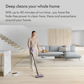 Model using Dyson V8 Advanced Cordless Stick Vacuum Cleaner in a bright open-plan living room with sofa and staircase visible.