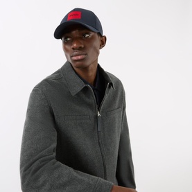 Model with Hugo Men-X Logo Baseball Cap in dark blue, six-panel design with red logo detail, styled with grey zip-up jacket