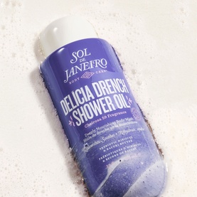 Sol De Janeiro Delicia Drench Shower Oil in purple bottle, shown surrounded by white foam lather