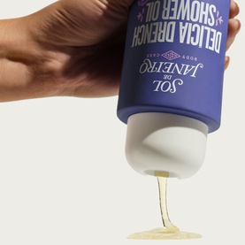 Sol De Janeiro Delicia Drench Shower Oil being squeezed from bottle, dispensing golden oil, purple packaging visible