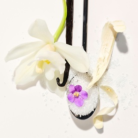 Sol De Janeiro Delicia Drench Shower Oil lifestyle shot with vanilla pods, white orchid flower, and a spoon of white crystals with violet flower