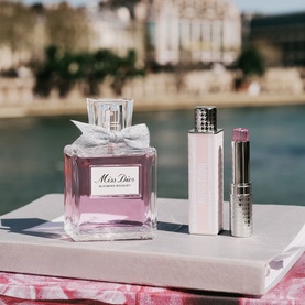 Dior Miss Dior Blooming Bouquet EDT bottle styled alongside a pink lipstick and lip gloss on a marble surface with Parisian backdrop.